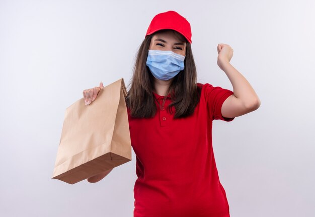 Freudige junge Lieferfrau, die rotes T-Shirt in der roten Kappe trägt, die ein Paket auf isolierter weißer Wand hält