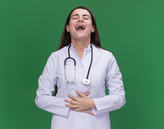 Freudige junge Ärztin, die medizinische Robe mit Stethoskop trägt, legt Hände auf Bauch und steht mit geschlossenen Augen auf Grün