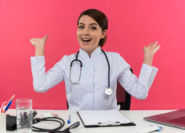 Freudige junge Ärztin, die medizinische Robe mit Stethoskop trägt, sitzt am Schreibtisch, der am Computer mit medizinischen Werkzeugen arbeitet, verbreitet Hände auf isolierter rosa Wand mit Kopienraum