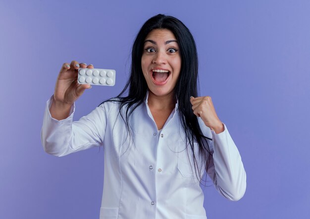 Freudige junge Ärztin, die medizinische Robe trägt, die Packung der medizinischen Tabletten zeigt und Faust ballt