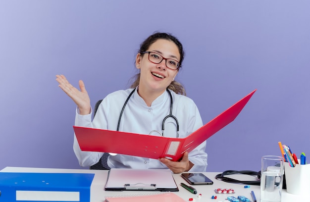 Freudige junge Ärztin, die medizinische Robe und Stethoskop trägt, sitzt am Schreibtisch mit medizinischen Werkzeugen, die Ordner suchen, der leere Hand lokalisiert zeigt