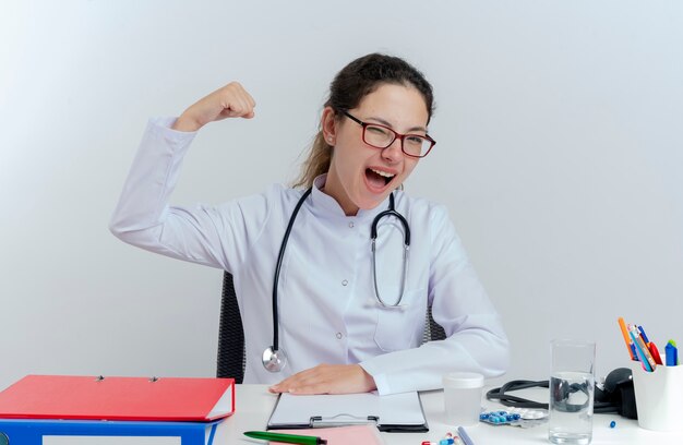 Freudige junge Ärztin, die medizinische Robe und Stethoskop und Brille trägt, die am Schreibtisch mit medizinischen Werkzeugen sitzen, die zwinkern, starke Geste lokalisiert tun