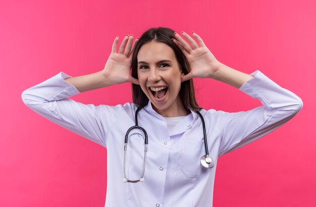 Freudige junge Ärztin, die medizinisches Kleid des Stethoskops trägt, legte ihre Hände um Ohren auf isolierte rosa Wand