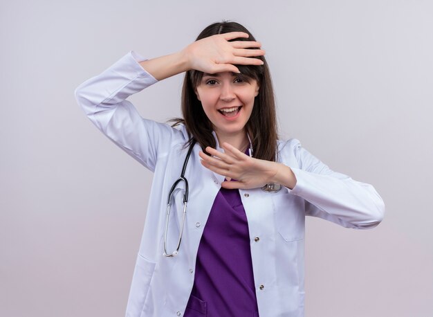 Freudige junge Ärztin im medizinischen Gewand mit Stethoskop erhebt die Hände auf lokalisiertem weißem Hintergrund