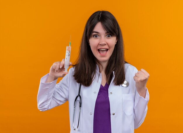 Freudige junge Ärztin im medizinischen Gewand mit Stethoskop hält Spritze und hält Faust auf lokalisiertem orange Hintergrund