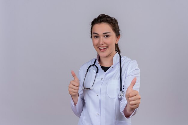 Freudige junge Ärztin im medizinischen Kleid mit Stethoskop zeigt ihren Daumen oben auf der weißen Wand