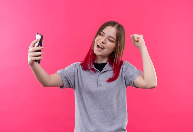 Freudige junge schöne Frau, die graues T-Shirt Takin Selfie trägt, das ja Geste auf isolierter rosa Wand zeigt