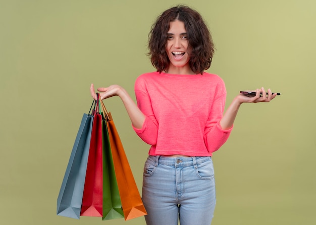 Freudige junge schöne Frau, die Kartontaschen und Handy auf isolierter grüner Wand hält