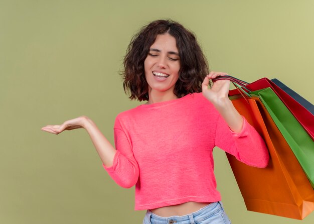 Freudige junge schöne Frau, die Papiertüten hält und leere Hand auf isolierter grüner Wand zeigt