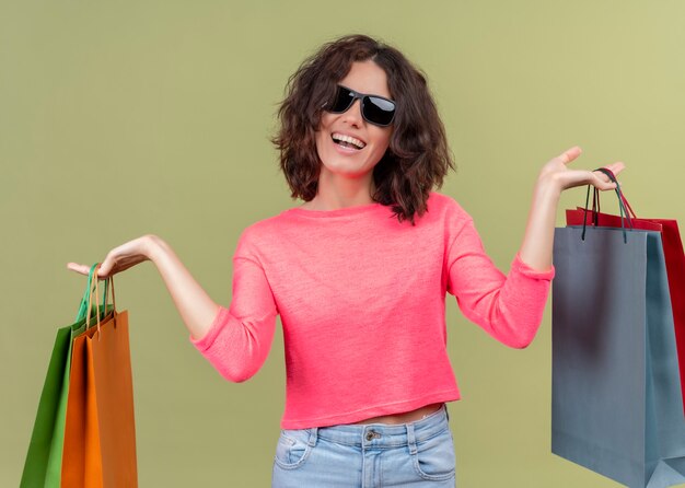 Freudige junge schöne Frau, die Sonnenbrille trägt und Kartontaschen auf isolierter grüner Wand hält