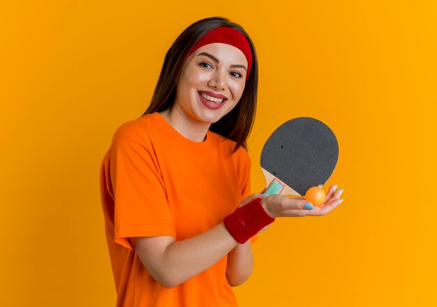 Freudige junge sportliche Frau, die Stirnband und Armbänder trägt, die Tischtennisschläger und Ball suchen