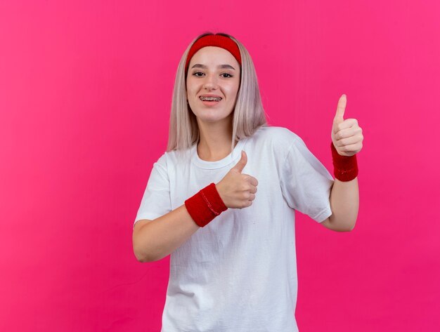 Freudige junge sportliche Frau mit Zahnspangen, die Stirnband und Armbänder Daumen hoch von zwei Händen tragen, die auf rosa Wand lokalisiert werden