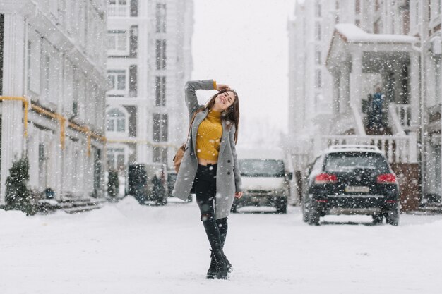 Freudige junge Winterfrau, die im Schneefall auf Straße in der Großstadt kühlt. Modisches Modell mit Rucksack, Lächeln mit geschlossenen Augen, Schneefall, Schneeflocken, Weihnachtsstimmung genießend.