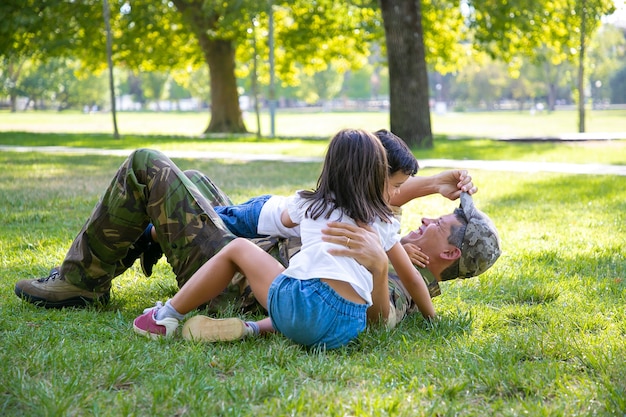 Freudige Kinder und ihr Vater liegen und spielen auf Gras im Park. Glückliches militärisches Vatertreffen mit Kindern nach Missionsreise. Familientreffen oder Rückkehr nach Hause Konzept