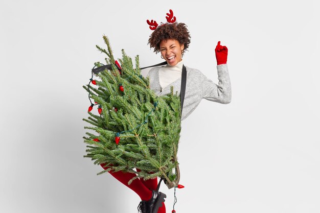 Freudige lockige Frau tanzt sorglos hat Spaß, bevor das Neujahrskonzert einen grünen Tannenbaum hält, als ob die Gitarre den Arm hebt und eine spielerische Stimmung hat, glücklich, allein zu Hause zu bleiben, trägt Rentierhörner. Winterferien