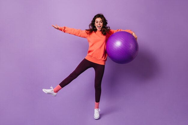Freudige lockige Sportlerin, die Spaß im lila Studio mit großem fitball hat
