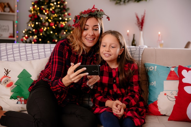 Freudige Mutter und Tochter, die Telefon betrachten, das auf Couch sitzt und Weihnachtszeit zu Hause genießt