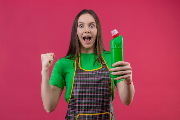 Freudige Reinigung der jungen Frau, die Uniform hält, die Reinigungsmittel hält, die ja Geste auf isolierter rosa Wand zeigt