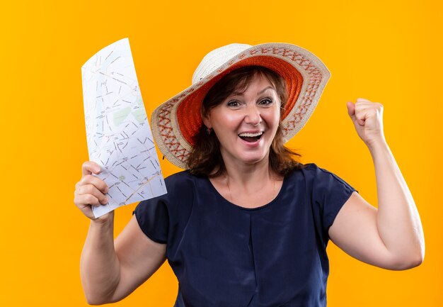 Freudige reisende Frau mittleren Alters im Hut, der Karte hält und Ja-Geste auf isolierter gelber Wand zeigt