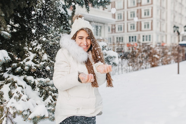 Freudige süße Frau, die Spaß mit Schneeflocken im Freien auf Tannenbaum voll mit Schnee hat. Junges charmantes Modell in der warmen Winterkleidung, die kaltes Schnee auf Straße genießt. Positivität ausdrücken, lächeln.