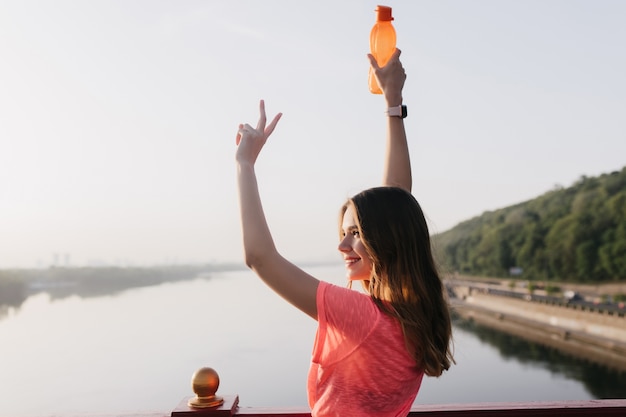 Freudige weiße Frau, die mit Lächeln aufwirft. Außenporträt des inspirierten sportlichen Mädchens mit Flasche.