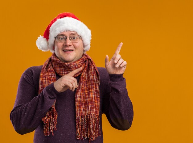 Freudiger erwachsener Mann, der Brille und Weihnachtsmütze mit Schal um den Hals trägt und Kamera betrachtet, die lokal auf orange Hintergrund mit Kopienraum zeigt