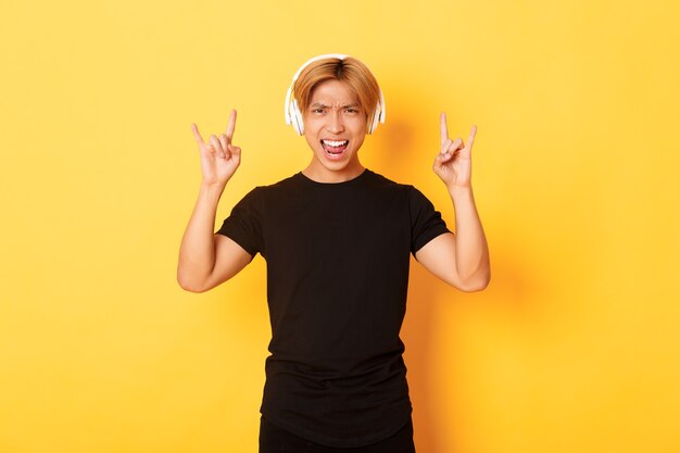 Freudiger hübscher asiatischer Kerl, der genießt, Musik in Kopfhörern zu hören, Rock'n'Roll-Geste zeigend, über gelber Wand stehend