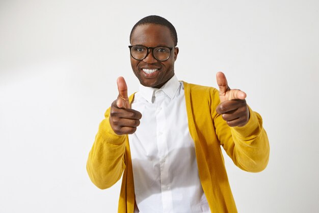 freudiger junger afroamerikanischer männlicher Hipster, der stilvolle Brillen und Strickjacke trägt, die breit lächeln, Zeigefinger zeigen und positiven freundlichen Gesichtsausdruck haben