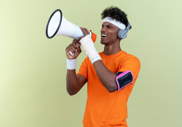 Freudiger junger afroamerikanischer sportlicher Mann, der Stirnband und Armband und Telefonarmband mit Kopfhörern trägt, spricht auf Lautsprecher lokalisiert auf grünem Hintergrund