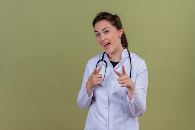 Freudiger junger Arzt, der medizinisches Kleid trägt, das Stethoskop trägt, zeigt auf grüne Wand voraus