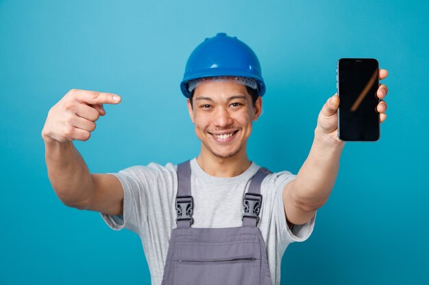 Freudiger junger Bauarbeiter, der Schutzhelm und Uniform trägt, streckt Handy in Richtung Kamera aus, die darauf zeigt