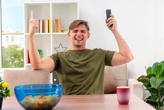 Freudiger junger blonder gutaussehender Mann sitzt am Tisch mit Schüssel der Chips und Tasse, die TV-Fernbedienung hält und Faust erhebt