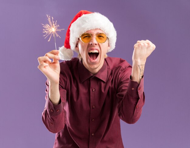 Freudiger junger blonder Mann, der Weihnachtsmütze und Brille hält, die Feiertagswunderkerze hält, die ja Geste lokalisiert auf lila Wand tut