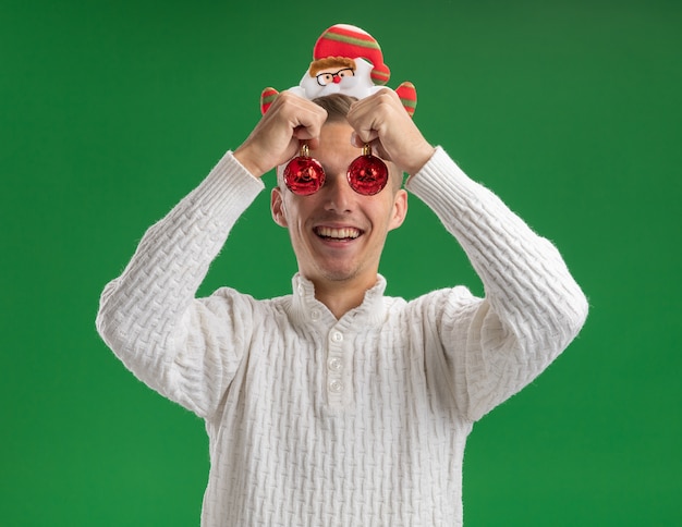 Freudiger junger gutaussehender Kerl, der Weihnachtsmann-Stirnband mit Brille trägt, die Weihnachtskugeln vor Augen lokalisiert auf grüner Wand hält