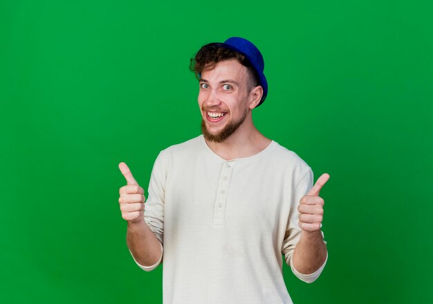 Freudiger junger gutaussehender slawischer Party-Typ, der Partyhut trägt, der Front zeigt, zeigt Daumen hoch lokalisiert auf grüner Wand mit Kopienraum