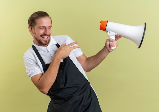 Freudiger junger hübscher Friseur, der Sprecher schaut und zur Seite zeigt, die auf olivgrün lokalisiert wird