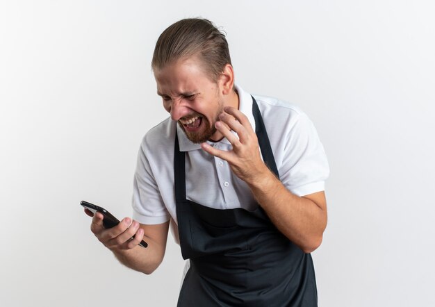 Freudiger junger hübscher Friseur, der Uniform hält und Handy hält, das Hand auf Luft lokalisiert auf Weiß hält