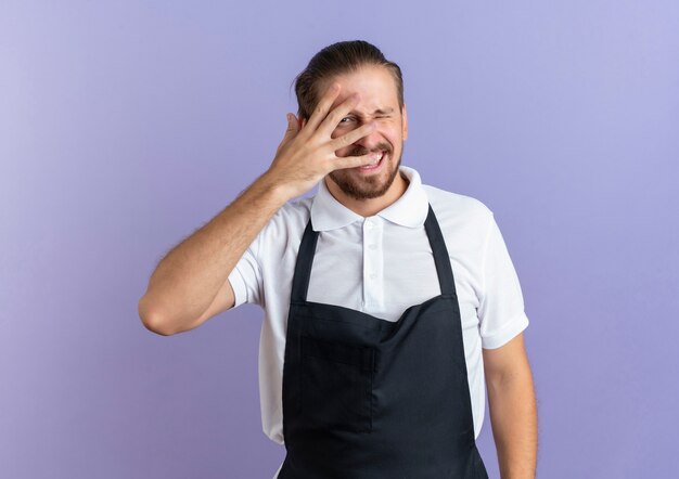 Freudiger junger hübscher Friseur, der Uniform trägt, die Hand auf Gesicht setzt, das durch Finger schaut und an Front, die auf Lila isoliert ist, zwinkert