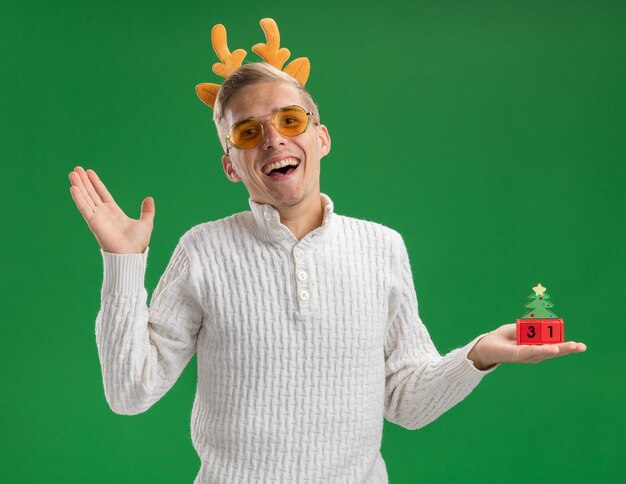 Kostenloses Foto freudiger junger hübscher kerl, der rentiergeweih-stirnband mit brille trägt, die weihnachtsbaumspielzeug mit datum hält, das leere hand lokalisiert auf grüner wand zeigt