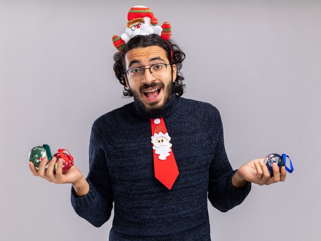 Freudiger junger hübscher Kerl, der Weihnachtskrawatte mit Haarbügel hält, der Weihnachtskugeln isoliert auf weißer Wand hält