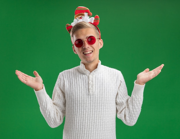 Freudiger junger hübscher Kerl, der Weihnachtsmann-Stirnband mit Brille trägt, die Kamera betrachtet, die leere Hände lokalisiert auf grünem Hintergrund zeigt