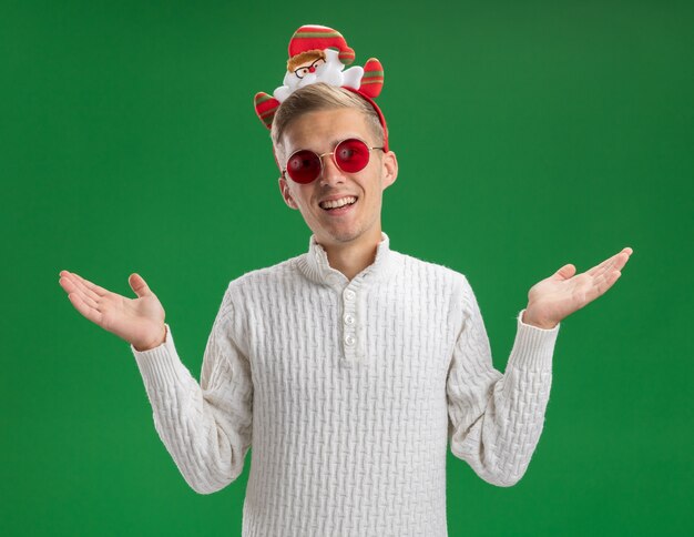 Freudiger junger hübscher Kerl, der Weihnachtsmann-Stirnband mit Brille trägt, die leere Hände lokalisiert auf grüner Wand zeigt