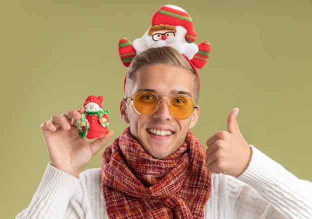 Freudiger junger hübscher Kerl, der Weihnachtsmann-Stirnband und Schal hält Schneemann-Weihnachtsverzierung, der Daumen oben auf olivgrüner Wand zeigt