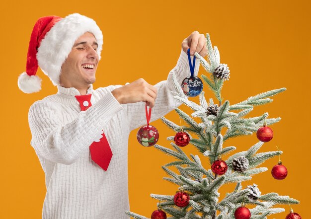 Freudiger junger hübscher Kerl, der Weihnachtsmütze und Weihnachtsmannkrawatte trägt, die nahe Weihnachtsbaum steht, der sie mit Weihnachtsballverzierungen verziert, die auf orange Wand lokalisiert werden