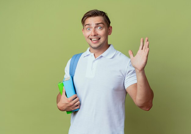 Freudiger junger hübscher männlicher Student, der Rückentasche hält Bücher hält und hallo lokalisiert auf olivgrünem Hintergrund gegeben wird