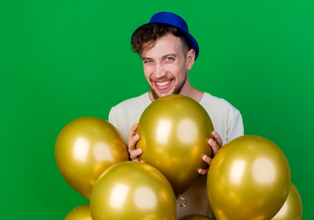 Freudiger junger hübscher slawischer Partei-Kerl, der Parteihut trägt, der hinter Luftballons steht und Kamera hält, der Ballon lokalisiert auf grünem Hintergrund hält