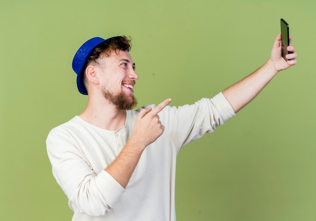 Freudiger junger hübscher slawischer Partei-Kerl, der Parteihut trägt, der selfie zeigt, das auf Handy lokalisiert auf olivgrünem Hintergrund zeigt