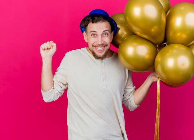 Freudiger junger hübscher slawischer Party-Typ, der Partyhut hält, der Ballons hält, die Front, die geballte Faust lokalisiert, lokalisiert auf rosa Wand betrachtet