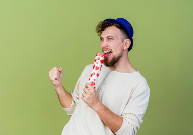 Freudiger junger hübscher slawischer Party-Typ, der Partyhut trägt, der gerade singend unter Verwendung von Konfettikanone als Mikrofon lokalisiert auf olivgrünem Hintergrund mit Kopienraum schaut