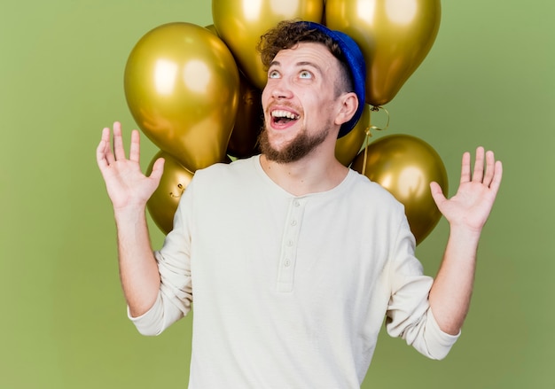 Freudiger junger hübscher slawischer Party-Typ, der Partyhut trägt, der vor Luftballons steht, die leere Hände zeigen, die lokalisiert auf olivgrünem Hintergrund suchen